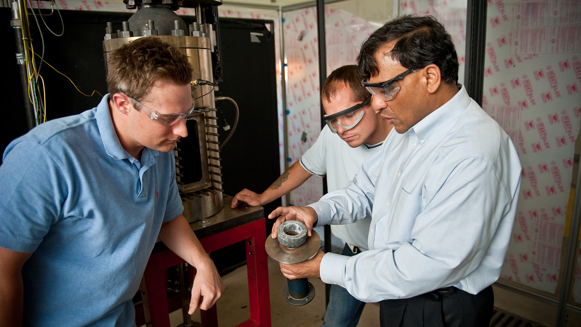 Dr. Ajay Agrawal shows a round piece of machinery to 2 students